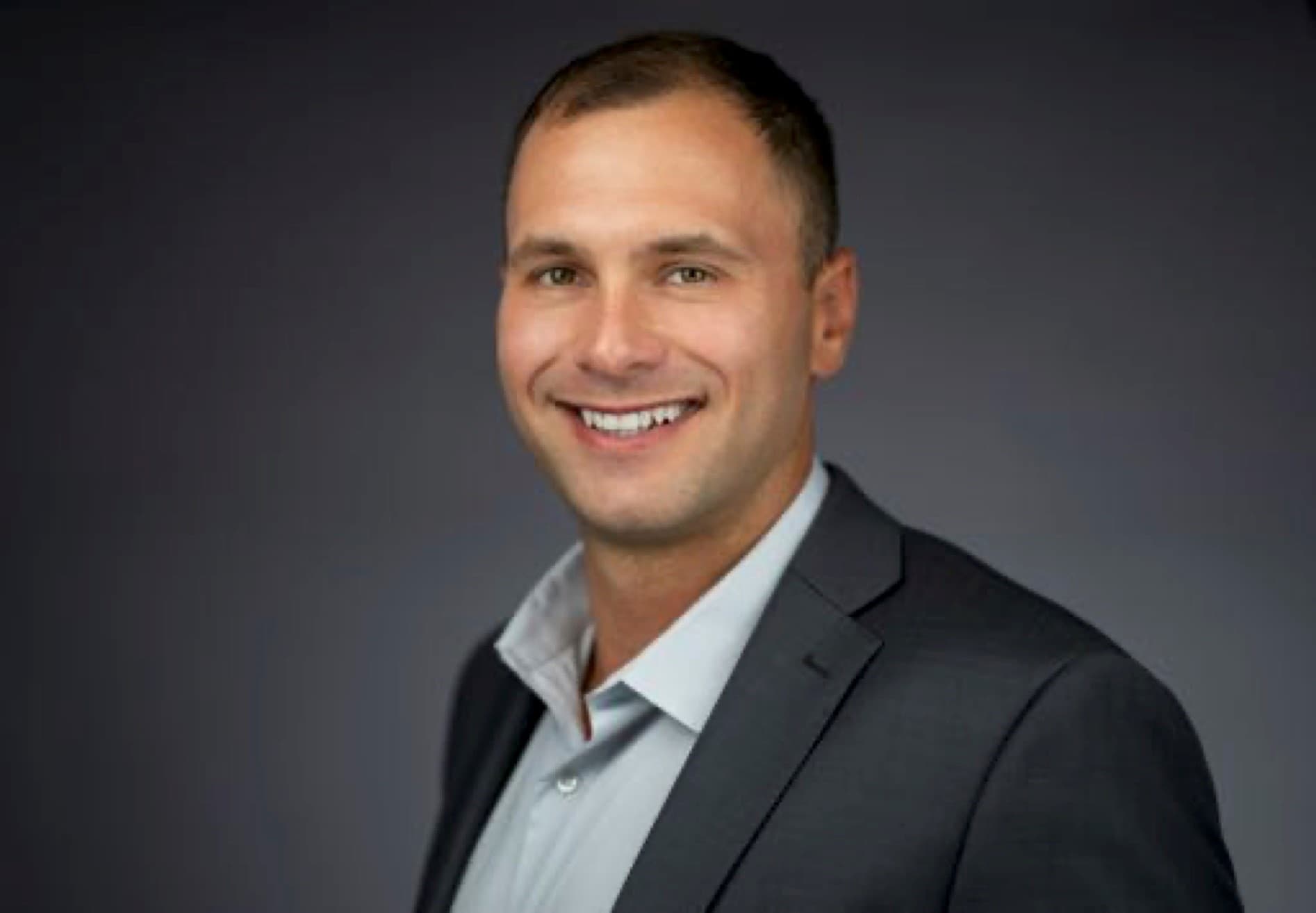 Smiling man in a grey suit jacket and light shirt, professional headshot against dark background.